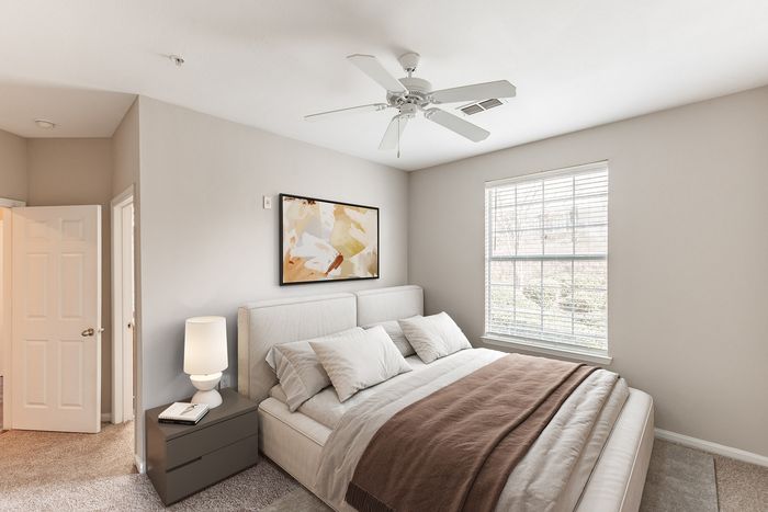 Bright and inviting bedroom featuring a large window, cozy bed with plush pillows, modern artwork, and a sleek nightstand with a stylish lamp.