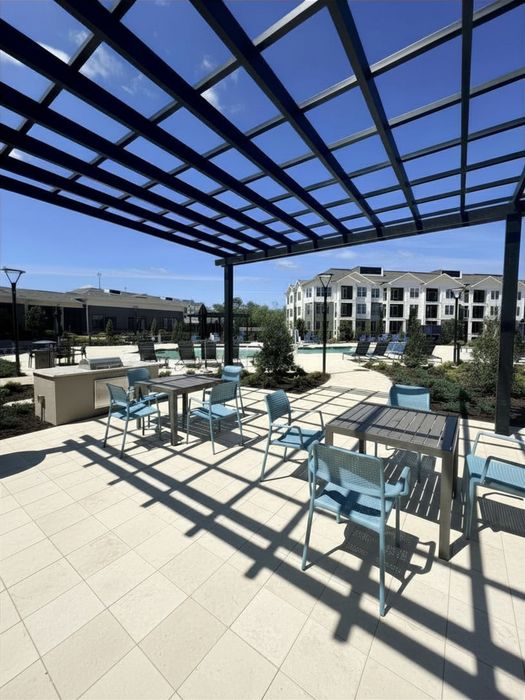 Modern apartment complex outdoor area featuring blue chairs and tables under a stylish pergola, with a scenic view of landscaped gardens and a spacious pool.