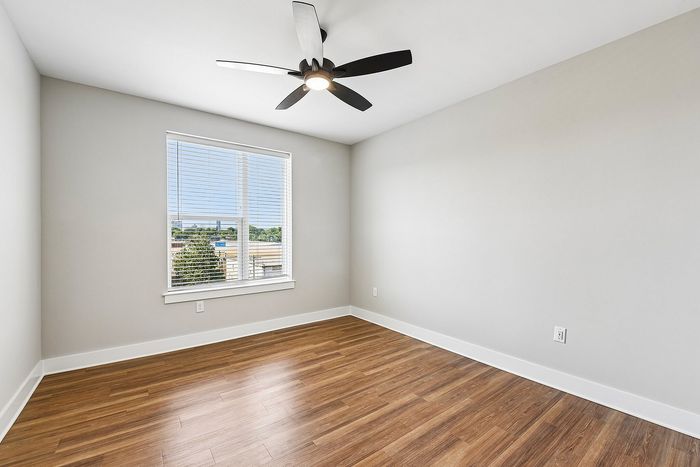 Bright and airy apartment room with elegant wood flooring, modern ceiling fan, and large window offering scenic views.