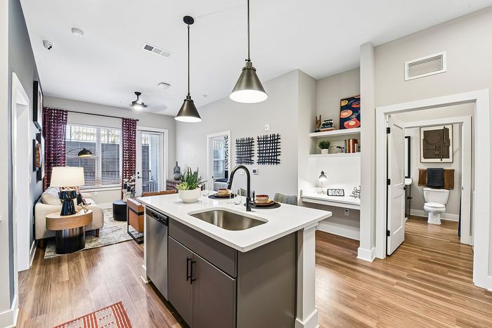 Modern apartment interior featuring an open-concept kitchen with sleek island, cozy living area, and decorated workspace, all highlighted by warm wood flooring and stylish lighting.