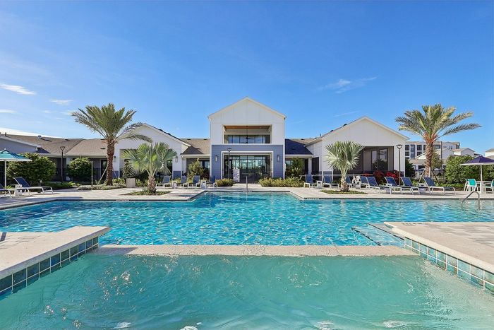 Luxurious apartment complex with a stunning outdoor pool area, surrounded by palm trees and stylish lounge chairs under a clear blue sky.