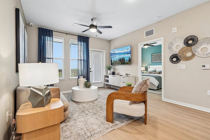 Bright living room with modern furnishings, wooden accents, and elegant wall art, leading to a cozy bedroom through an open doorway.