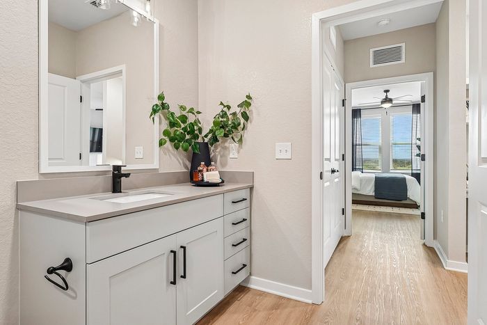 Bright modern bathroom with sleek vanity, accentuated by lush green plant, leading to a cozy, naturally lit bedroom with large windows.
