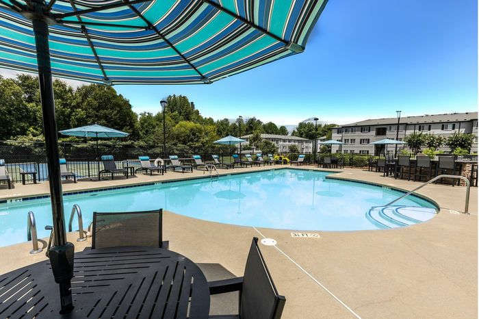 Luxurious apartment complex featuring a pristine outdoor swimming pool with stylish lounge chairs and elegant umbrellas, surrounded by lush greenery and modern buildings.