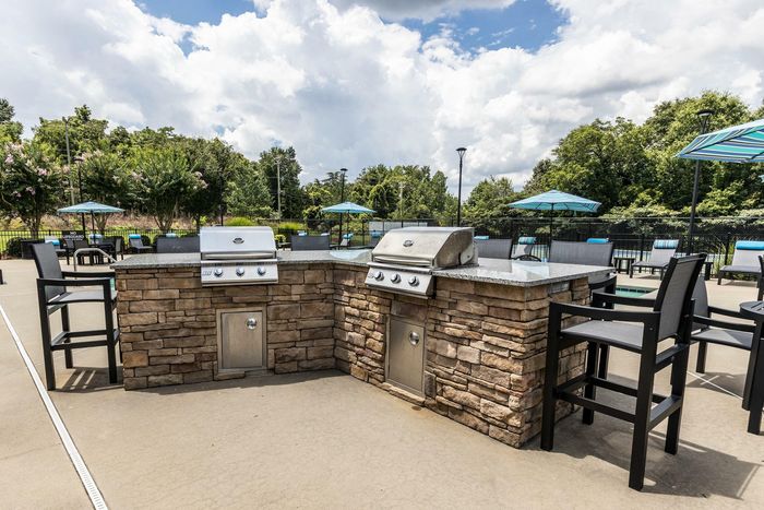 Inviting outdoor space featuring a modern stone grill area with high-top seating, surrounded by lush greenery and vibrant blue umbrellas.