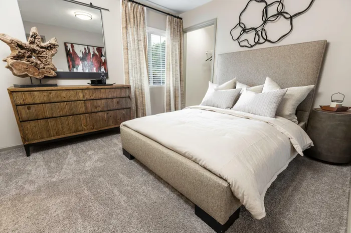 Elegant bedroom with plush carpeting, featuring a modern beige bed, stylish wooden dresser, and artistic wall decor.