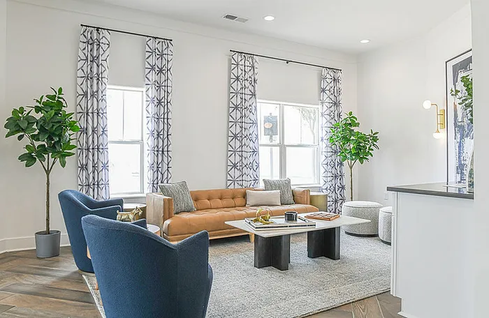 Modern living room with stylish decor featuring a plush tan sofa, elegant blue chairs, and abundant natural light from large windows with patterned curtains.