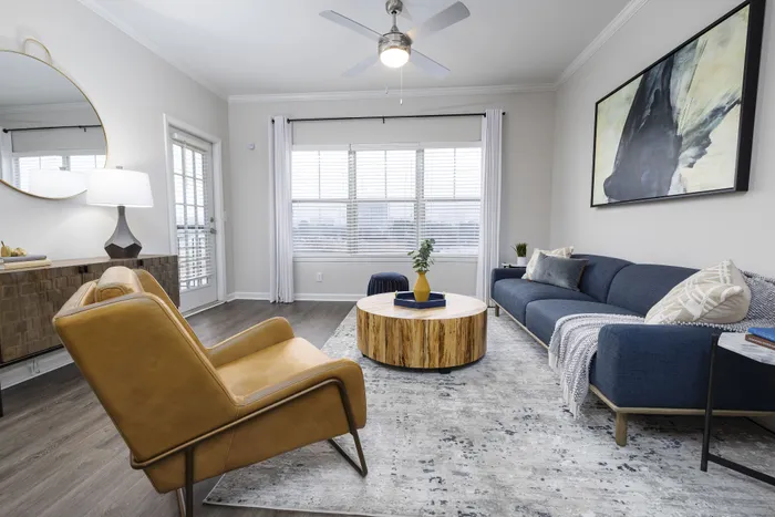 Sophisticated living room with elegant furniture, featuring a modern blue sofa, stylish mustard armchair, and a chic round wooden coffee table, beautifully lit by natural light from large windows.