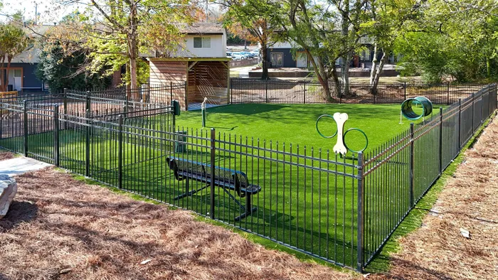 Modern fenced dog park with lush green turf, agility equipment, and comfortable seating surrounded by trees.