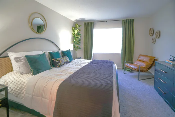 Chic bedroom with cozy bedding, vibrant pillows, and stylish decor, featuring a lush plant and modern accent chair in a bright, inviting space.