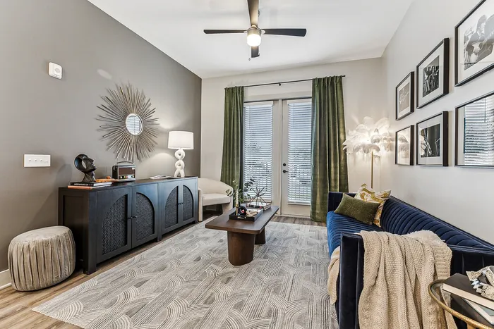 Stylish living room with modern decor, featuring a plush blue sofa, elegant wall art, and a chic sunburst mirror. Large windows with green curtains let in natural light.