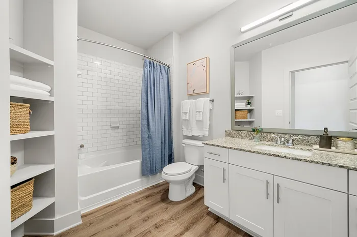 Elegant and modern bathroom with white cabinetry, granite countertop, and subway tile shower. Features a soothing blue shower curtain, open shelving with chic wicker baskets, and sleek wood flooring.
