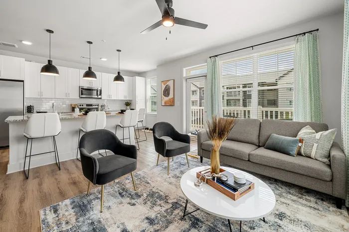 Modern open-concept living room and kitchen with elegant gray and black furniture, sleek pendant lighting, and large sunlit windows.