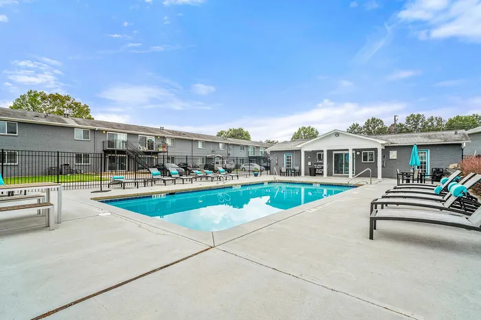 Inviting apartment complex with a sparkling swimming pool surrounded by stylish lounge chairs, perfect for relaxation under a vibrant blue sky.