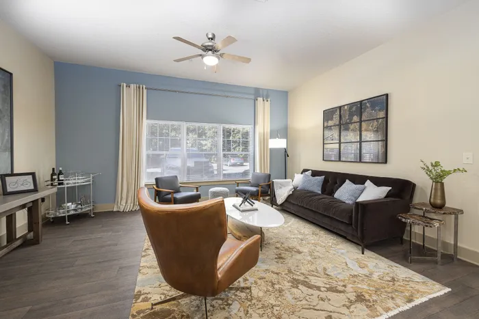 Chic and inviting living room featuring a plush dark sofa, stylish armchairs, and a modern coffee table. Large windows offer ample natural light, complemented by elegant decor and a sophisticated ceiling fan.