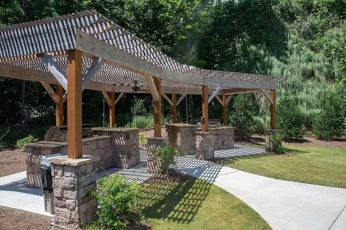 Charming outdoor BBQ area with stone counters and wooden pergola, set amidst lush greenery and inviting landscaped paths.