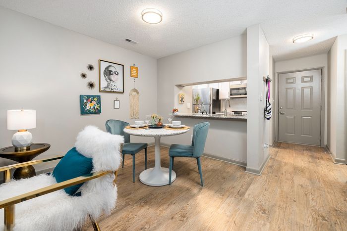 Chic apartment dining area showcasing a cozy round table set for two, elegant teal chairs, and modern artwork. Bright, open-plan layout with a view into a sleek kitchen featuring stainless steel appliances.