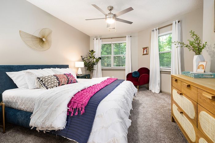 Bright and inviting bedroom with elegant decor, featuring a plush bed with colorful accents, modern ceiling fan, and abundant natural light from large windows.