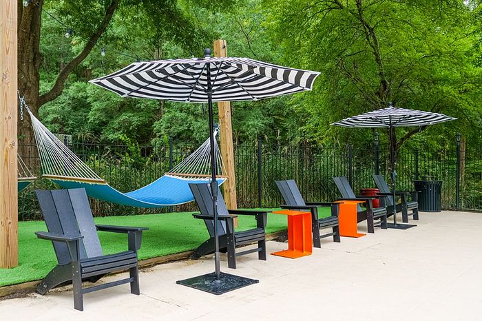 Inviting outdoor space with cozy hammocks, stylish black chairs, vibrant orange tables, and striped umbrellas, surrounded by lush greenery.