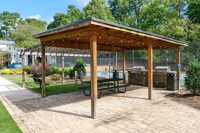 Charming outdoor area with a wooden pergola, picnic tables, and built-in grills beside lush greenery and a tennis court.