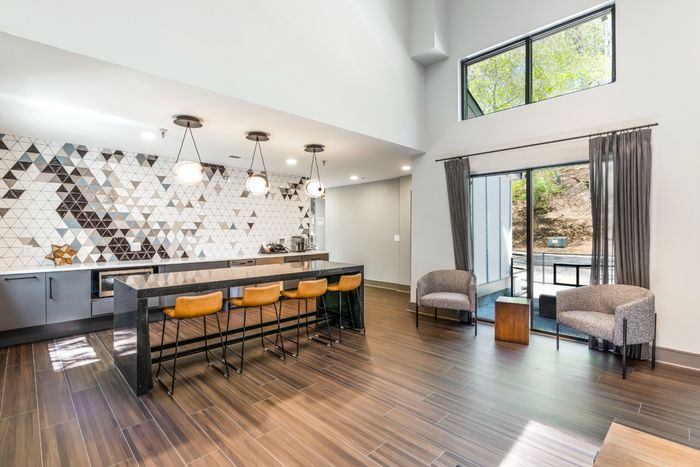 Modern kitchen and lounge area featuring sleek countertops, stylish geometric tile backsplash, wooden bar stools, and a spacious open layout with large windows.