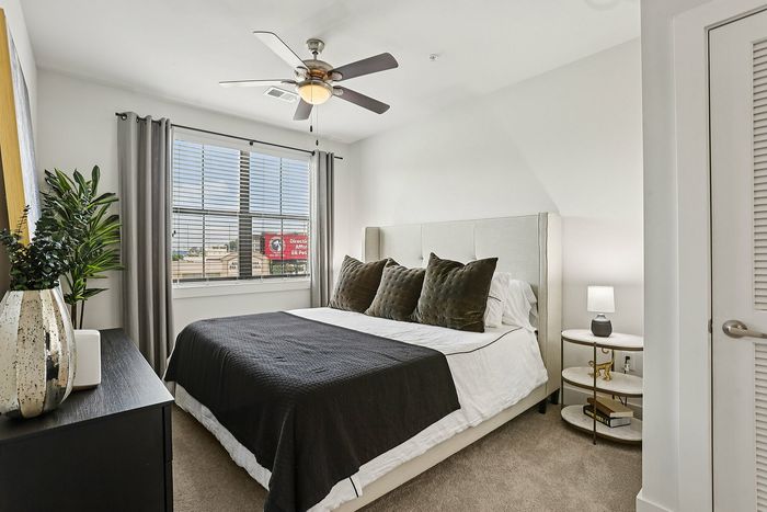 Elegant bedroom featuring a plush bed with dark cushions, modern ceiling fan, and natural light from large windows.