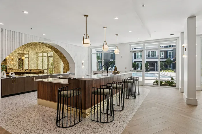 Luxurious apartment community lounge featuring a sleek kitchen island, modern bar seating, and poolside views through expansive windows.