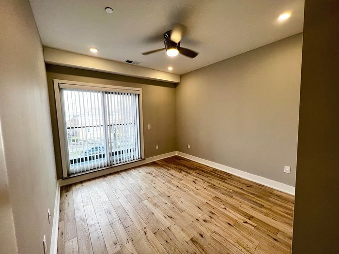 Inviting empty room with wooden flooring, ceiling fan, and large window with blinds.