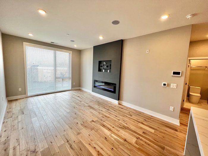 Bright and airy living room with wooden floors and modern fireplace, featuring a large window with blinds.