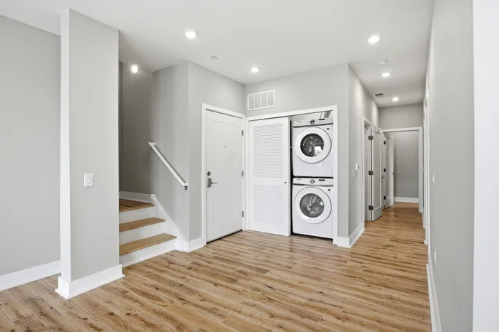 Modern apartment hallway featuring sleek wood flooring, convenient in-unit laundry with stacked washer and dryer, and bright recessed lighting.