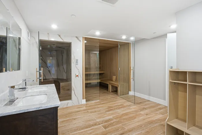 Luxurious bathroom with elegant marble sinks, a modern sauna, and a spacious steam room, featuring sleek glass doors and warm wooden floors.