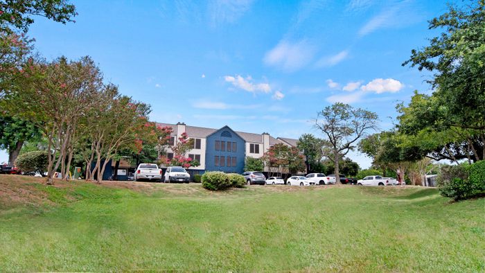 Charming apartment complex with lush greenery and ample parking under a bright blue sky.