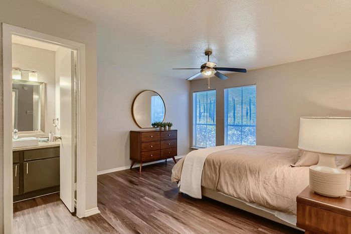 Inviting bedroom with elegant hardwood floors, modern ceiling fan, and ensuite bathroom; natural light streams through large windows for a bright, airy feel.