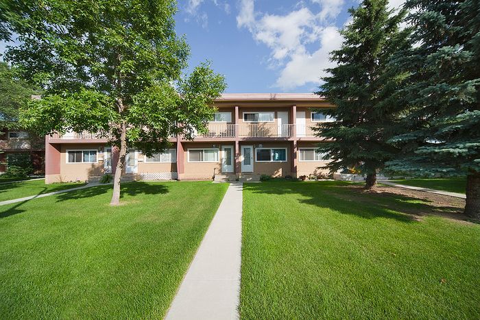 Charming apartment complex featuring lush green lawns and a scenic walkway leading to welcoming entrances, nestled among mature trees under a vibrant blue sky.