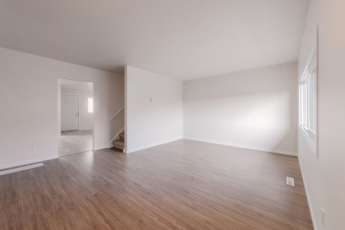 Bright and spacious living area with elegant wooden flooring and large windows, leading to a staircase and open doorway.