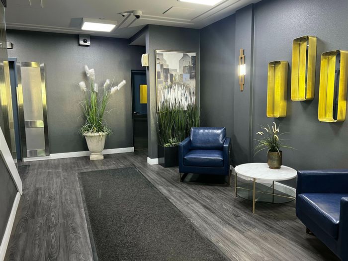 Elegant apartment lobby with modern gray walls, plush blue chairs, decorative plants, and stylish lighting.