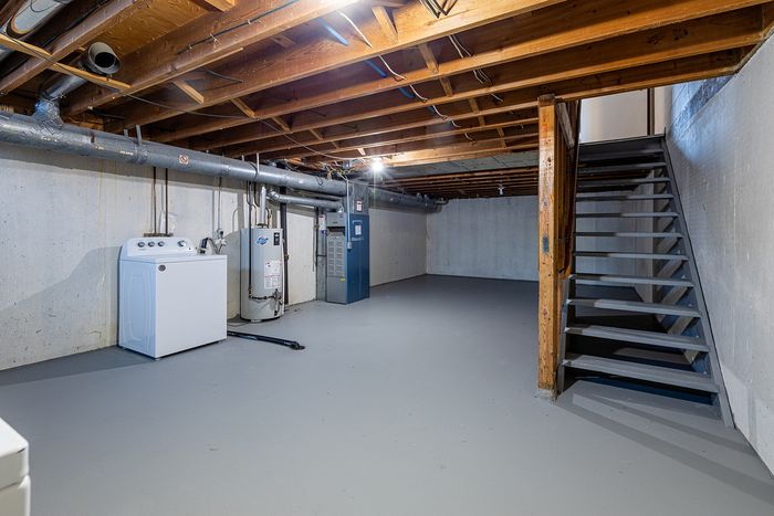 Spacious unfinished basement featuring exposed wooden beams, utility area with washer and water heater, and a sturdy staircase, ideal for customization.