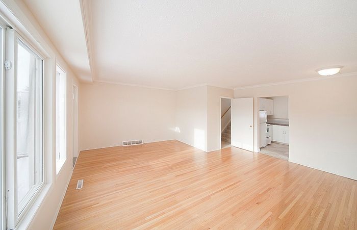 Bright and spacious empty living room with polished hardwood floors, large windows, and an open doorway leading to a modern kitchen.