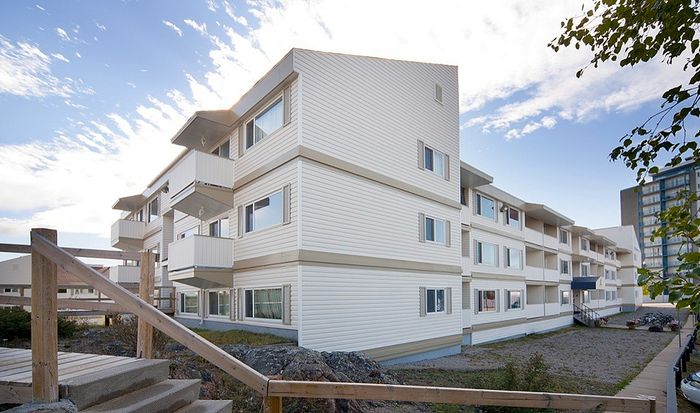 Modern multi-story apartment building with white siding, featuring spacious balconies and large windows, under a bright blue sky.