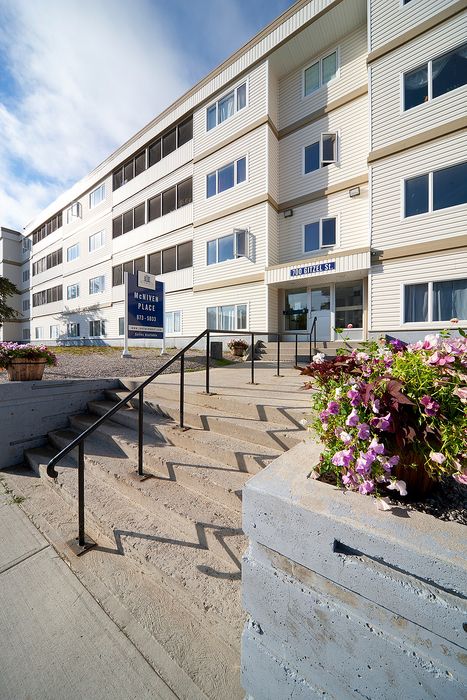 Inviting entrance of a modern apartment building featuring crisp white siding, beautifully maintained flower planters, and welcoming stairs leading up to McNiven Place.