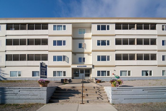 Charming multi-story apartment building with a welcoming entrance, surrounded by flower pots and clear blue skies.