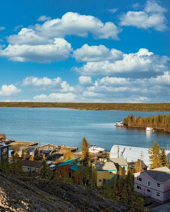 Scenic waterfront view with colorful houses, lush green trees, and serene lake under a vibrant blue sky with fluffy clouds.