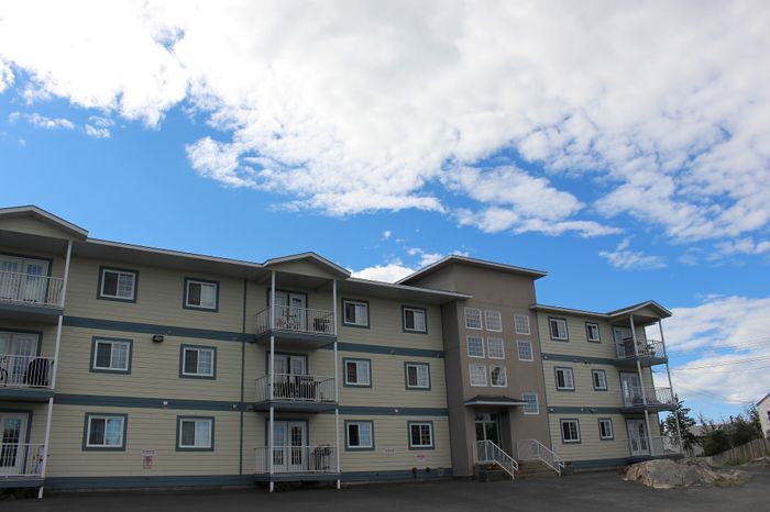 Modern three-story apartment building under a bright blue sky, featuring private balconies and ample sunlight.