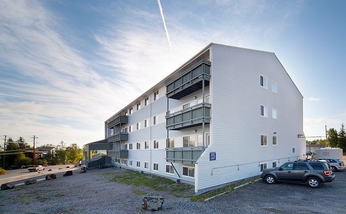Modern four-story apartment building with private balconies and ample parking set against a clear blue sky.