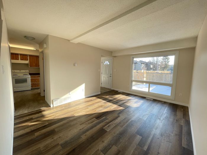 Bright living area with large window and open view, featuring stylish wood flooring, adjacent to a cozy kitchen with wooden cabinets.