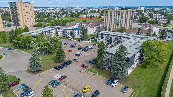 Aerial view of a vibrant apartment complex with well-maintained grounds, ample parking, and lush greenery, set against a backdrop of a bustling cityscape.