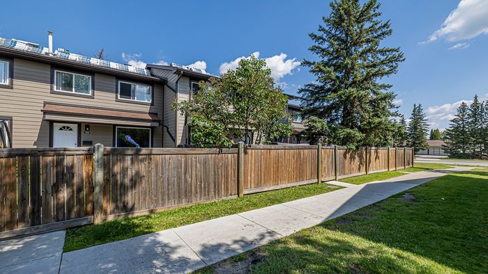 Charming townhouse with a modern exterior, nestled behind a wooden fence, surrounded by lush greenery and tall trees under a clear blue sky.