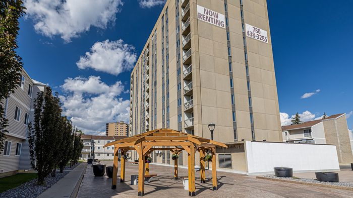 Modern high-rise apartment with a 'Now Renting' sign, featuring a vibrant courtyard gazebo surrounded by landscaped greenery under a clear blue sky.