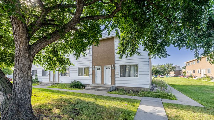 Charming townhouse exterior featuring a white facade, dual entry doors, and lush green surroundings under a large shade tree.