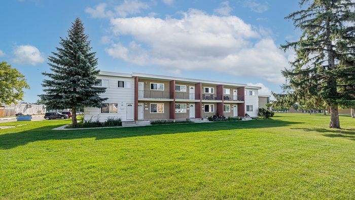 Charming apartment building with a spacious lawn, surrounded by lush trees and set against a bright blue sky.